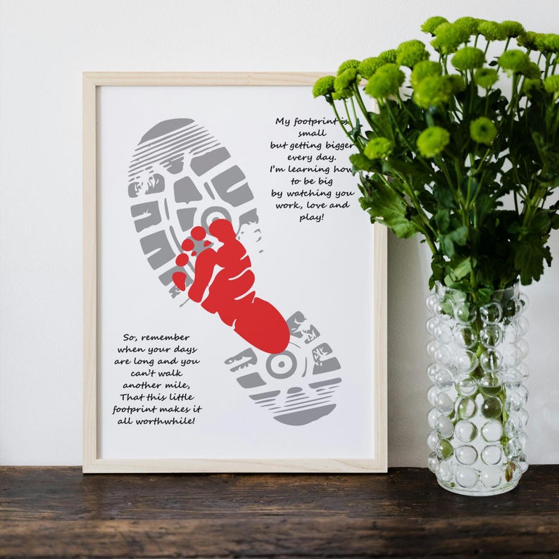 Op de afbeelding: Een ingelijste print met een grijs voetstappenontwerp en een rode babyvoet. De print bevat inspirerende tekst over groei en liefde. Een vaas met groene bloemen staat rechts. De lijst is van licht hout.
