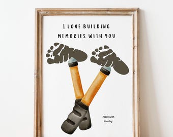 Op de afbeelding: Een ingelijst print met de tekst "Ik hou van het bouwen van herinneringen met jou" en twee handafdrukken die een paar werkschoenen vormen. De schoenen zijn oranje en zwart geverfd.
