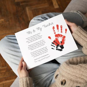 May include: A white card with the text "Me & My Teacher" and a poem. The card features a red handprint over a black handprint. The card is held by a person wearing a beige sweater and blue jeans.