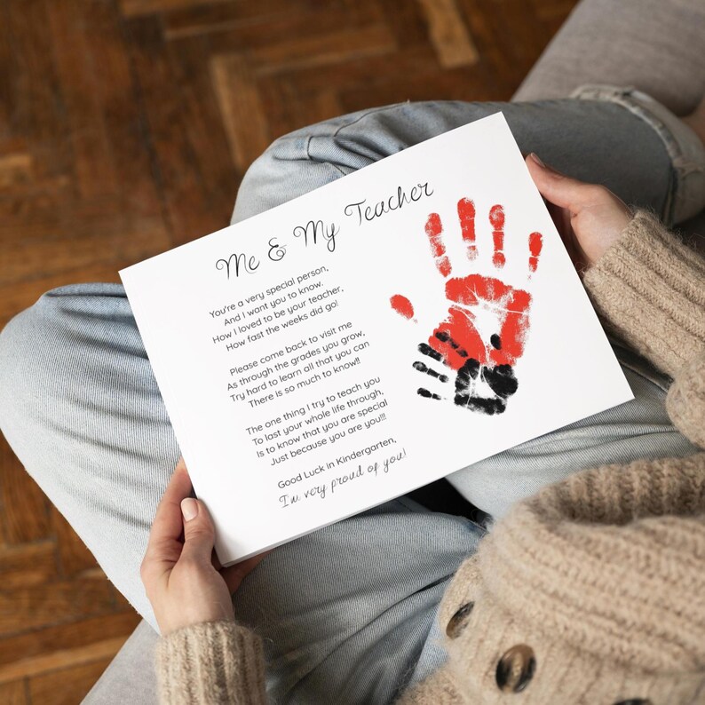 May include: A white card with the text "Me & My Teacher" and a poem. The card features a red handprint over a black handprint. The card is held by a person wearing a beige sweater and blue jeans.