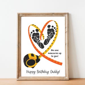 May include: A framed print featuring a heart shape made of a yellow and orange measuring tape, with two black baby footprints inside. The print includes the text "No one measures up to you!" and "Happy Birthday Daddy!"