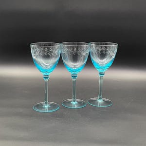 May include: Three stemmed wine glasses with clear bowls and teal blue stems and bases. Each glass features an etched leaf design. The glasses are arranged in a row against a dark background.