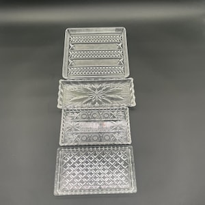 Op de afbeelding: Vier heldere rechthoekige glazen trays met diverse reliëfpatronen. De trays zijn gestapeld op een donkere achtergrond. De patronen omvatten geometrische vormen en stervormige ontwerpen. Deze trays zijn geschikt voor serveren of presenteren.