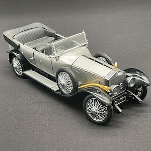 Può includere: Un modellino di auto decappottabile in stile vintage color argento e nero, caratterizzato da una griglia anteriore prominente, ruote a raggi, un ornamento dorato sul cofano e interni aperti dettagliati con sedili a panchetta e parafanghi neri.