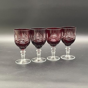 May include: Four ruby red wine glasses with clear stems and bases. Each glass features a detailed, etched design radiating from the center. The glasses are arranged in a row against a dark background, showcasing their elegant form and color.