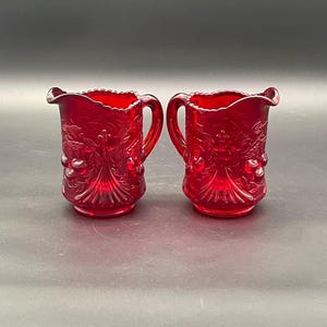 Pode incluir: Duas jarras de vidro vermelho rubi com desenhos florais e de bagas ornamentados. Cada jarra tem uma alça e uma borda com babados. As jarras são exibidas em uma superfície cinza escuro.