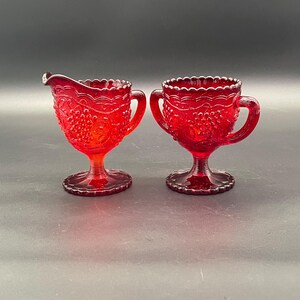 May include: Two ruby red glass serving pieces. The creamer has a spout and handle, while the sugar bowl has two handles. Both pieces have a grape design and scalloped edges. The pieces are on a pedestal base.