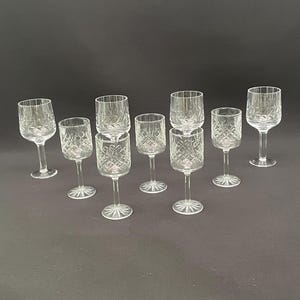 Op de afbeelding: Een set van acht heldere glazen op voet met een geslepen diamant- en sterpatroon op de kelken. Elk glas heeft een slanke steel en een ronde voet met een stralend sterontwerp, gegroepeerd op een donker oppervlak.