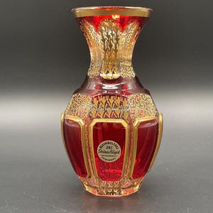 May include: A decorative red and gold crystal vase. The vase features a gold rim and ornate gold patterns. The label on the vase reads "Bleikristall 24% Steiner Vogel Handarbeit Germany."
