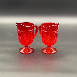 May include: Two red glass cream jugs with a detailed, textured pattern. Each jug has a handle and a small spout. The jugs are on a pedestal base and are displayed against a dark background.