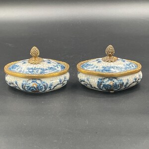 Könnte beinhalten: Zwei ovale Schmuckdosen aus Keramik mit Deckel. Die Dosen haben ein blau-weißes Blumenmuster mit goldfarbenen Rändern und eine goldfarbene, zapfenförmige Verzierung auf dem Deckel. Sie werden auf einer dunklen Oberfläche präsentiert.