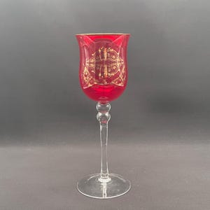 Könnte beinhalten: Ein hohes Stielglas mit einer tiefroten Kuppa, die mit komplexen goldenen Filigranmustern verziert ist und einen goldenen Rand aufweist. Der Stiel besteht aus klarem Glas mit einem kleinen kugelförmigen Knoten, auf einem runden Glasfuß.