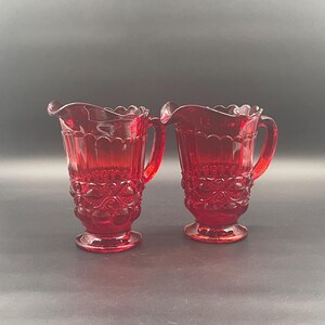 Pode incluir: Duas jarras de vidro vermelho rubi com bordas onduladas e detalhes florais ornamentados. Cada jarra tem uma alça e assenta sobre uma base de pedestal. As jarras de vidro transparente são adequadas para servir bebidas.