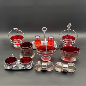 Könnte beinhalten: Eine Sammlung von Vintage-Servierschalen und Zubehör aus rubinrotem Glas. Enthält Salz- und Pfefferstreuer, Schalen, ein Sahne- und Zuckerdosen-Set und Serviettenringe. Die Teile sind auf einer dunklen Oberfläche angeordnet.