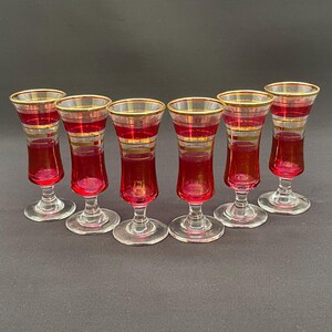 Peut inclure: Six verres à liqueur vintage, avec une coupe rouge rubis et un pied transparent. Chaque verre est orné d'une bordure dorée et de bandes dorées. Les verres sont disposés en demi-cercle sur un fond sombre.
