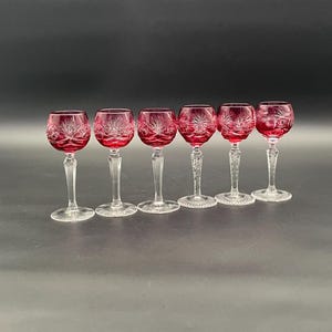 Könnte beinhalten: Sechs Weingläser mit rubinroten Kelchen und klaren Stielen. Die Kelche weisen ein Sternmuster auf. Die Gläser sind in einer Reihe vor einem dunklen Hintergrund angeordnet.