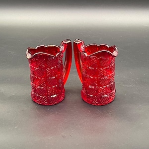 Könnte beinhalten: Zwei rubinrote Glaskrüge mit strukturierten Diamant- und Blumenmustern. Jeder Krug hat einen Henkel und einen gewellten Rand. Die Krüge werden vor einem dunklen Hintergrund präsentiert.