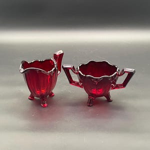 Può includere: Un set di lattiera e zuccheriera in vetro rosso. La lattiera ha un beccuccio e un manico, mentre la zuccheriera ha due manici. Entrambi i pezzi hanno un bordo smerlato e poggiano su tre piedini. Il set è realizzato in vetro rosso traslucido.