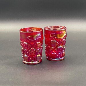 Puede incluir: Dos vasos de color rojo rubí con un patrón geométrico texturizado. Los vasos tienen una forma corta y cilíndrica y un acabado ligeramente iridiscente. La cristalería es probablemente vintage y adecuada para servir bebidas.