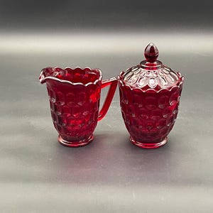 May include: A ruby red glass creamer and covered sugar bowl set. The creamer has a handle and a scalloped rim. The sugar bowl has a lid with a pointed finial. Both pieces have a textured, patterned design.