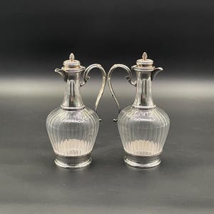 May include: Two silver and glass pitchers with a classic design. Each pitcher has a clear, ribbed glass body, a silver handle, and a silver lid. The pitchers are displayed against a dark background.