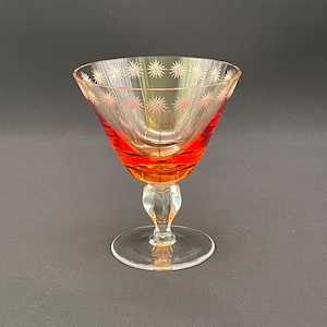 May include: A clear glass coupe with a vibrant orange-red bowl featuring a band of etched starburst patterns around the rim. The glass has a short, clear stem and a circular base, showcasing a distinct transition from the colored bowl to the clear stem.