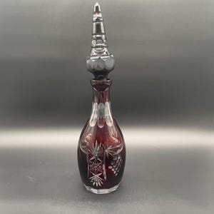 Könnte beinhalten: Eine rubinrote Glas-Dekantierkaraffe mit einem hohen, facettierten Verschluss. Die Karaffe weist geätzte Designs von Blättern und Trauben auf. Der Verschluss ist dunkelgrau und hat ein geometrisches Design. Die Karaffe ist etwa 30 cm hoch.