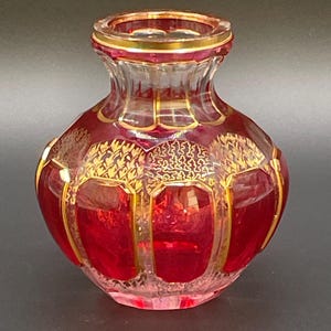 Könnte beinhalten: Kleine, dekorative Glasvase mit rundem Körper und schmalem Hals. Die Vase ist tiefrot mit goldfarbenen Akzenten und Verzierungen. Die goldfarbene Verzierung umrandet die Paneele und den Rand der Vase.
