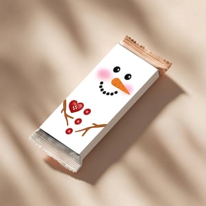 May include: A white candy bar wrapper featuring a snowman design. The snowman has black eyes, a carrot nose, and a smiling mouth. Pink blush accents the cheeks. A red heart and buttons complete the design. The wrapper is partially open.