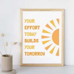 May include: A framed print with the text "YOUR EFFORT TODAY BUILDS YOUR TOMORROW" in orange. A stylized orange and yellow sun graphic is on the right. The frame is light brown, and a beige vase with dried flowers is in the foreground.