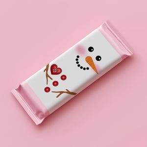May include: A pink and white candy bar wrapper featuring a snowman design. The snowman has black eyes, a carrot nose, and a coal mouth. Red heart and button accents complete the festive look. The wrapper is on a pink background.