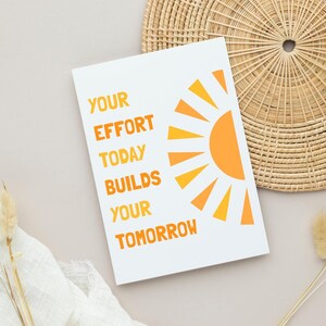 May include: White card with the text "YOUR EFFORT TODAY BUILDS YOUR TOMORROW" in orange. A stylized orange sun graphic is on the right. The card is on a neutral surface with a woven basket and dried plants.