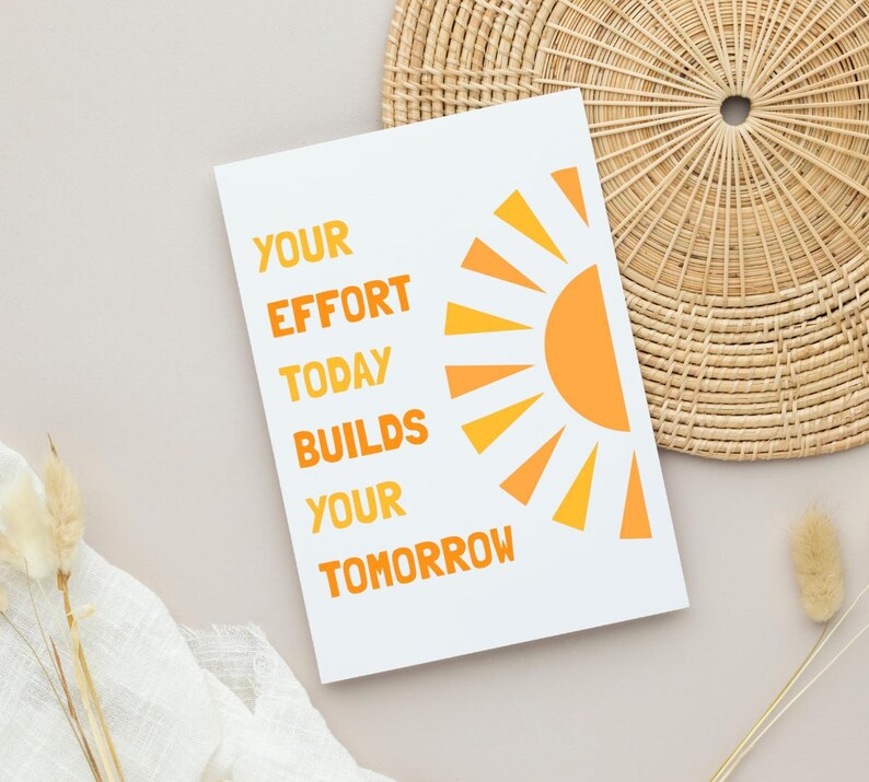 May include: White card with the text "YOUR EFFORT TODAY BUILDS YOUR TOMORROW" in orange. A stylized orange sun graphic is on the right. The card is on a neutral surface with a woven basket and dried plants.