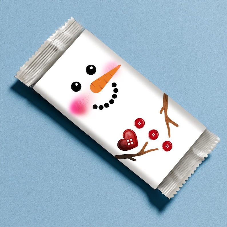 May include: A white candy bar wrapper featuring a snowman face with black eyes, an orange carrot nose, and a black dotted smile. Pink blush accents the cheeks. Red heart and button details complete the design. The wrapper is sealed with a silver foil edge.