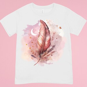 Op de afbeelding: Wit T-shirt met een aquarelontwerp van een roze en bruine veer met een halve maan en sterren op de achtergrond.