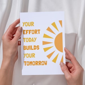 May include: A white card with the text "YOUR EFFORT TODAY BUILDS YOUR TOMORROW" in orange. A stylized orange sun graphic is on the right side of the card. The card is held by two hands.