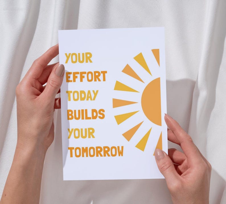 May include: A white card with the text "YOUR EFFORT TODAY BUILDS YOUR TOMORROW" in orange. A stylized orange sun graphic is on the right side of the card. The card is held by two hands.