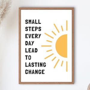 May include: A framed print with the words "SMALL STEPS EVERY DAY LEAD TO LASTING CHANGE" in black text. A yellow sun graphic is on the right. The frame is a light brown color. The background is white.