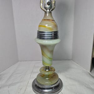 May include: A table lamp featuring a swirled glass body with cream, yellow, and green tones. The lamp has a silver-colored metal base and a matching silver-colored band around the center. A light bulb socket sits at the top with metal hardware.