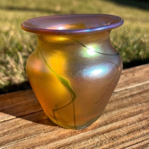 May include: A small, iridescent glass vase with a rounded body and a flared lip. The vase has a golden yellow base with green and blue iridescent streaks.