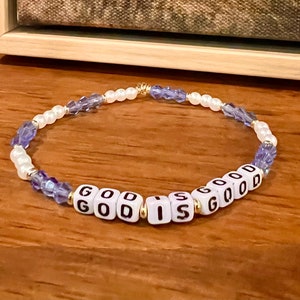 May include: A beaded bracelet with the words "GOD IS GOOD" spelled out in white letter beads. The bracelet also features white pearl beads, purple crystal beads, and gold accents. The bracelet is on a wooden surface.