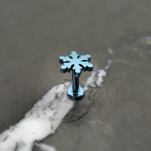 May include: A light blue snowflake-shaped lip piercing stud. The stud features a detailed snowflake design and a matching blue post and base. The jewelry is set against a mottled gray background.