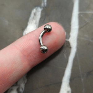 May include: A curved, silver-colored eyebrow piercing with two small, round balls at each end. The jewelry is made of a shiny, metallic material and is shown against a neutral background.