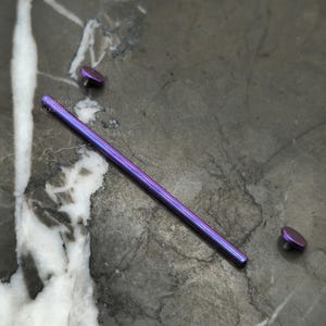 May include: A purple titanium barbell with two end balls. The barbell is lying on a gray marble surface.