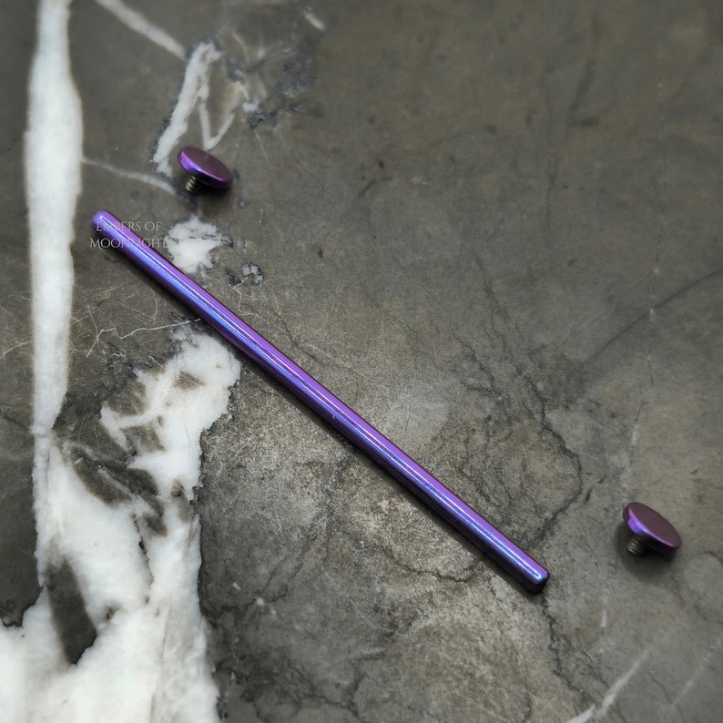 May include: A purple titanium barbell with two end balls. The barbell is lying on a gray marble surface.