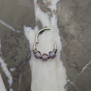 May include: A silver-colored hinged nose ring with three frosted lavender beads. The beads are strung on a wire that wraps around the ring's base. The ring is open and ready to be clasped.