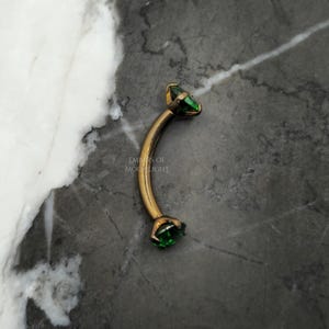 May include: Gold body jewelry with a curved barbell shape and two green gem accents.  The jewelry is on a gray background.