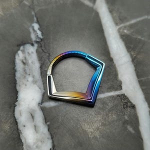 May include: A geometric, hinged ring with a rainbow iridescent finish. The ring has a pentagon shape with a curved top and a sharp, angular bottom. The colors transition from gold to blue, creating a vibrant, eye-catching accessory.