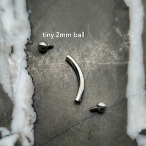 May include: A silver-toned curved barbell body jewelry piece consisting of a smooth, arched metal bar and two detachable ends, each featuring a tiny 2mm ball with a threaded post. The components are displayed separately on a dark gray surface.