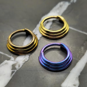 May include: Three metal hoop earrings in gold and iridescent blue-purple hues. Each earring features a layered design, with multiple stacked rings creating a textured appearance. The earrings are displayed on a marbled surface.